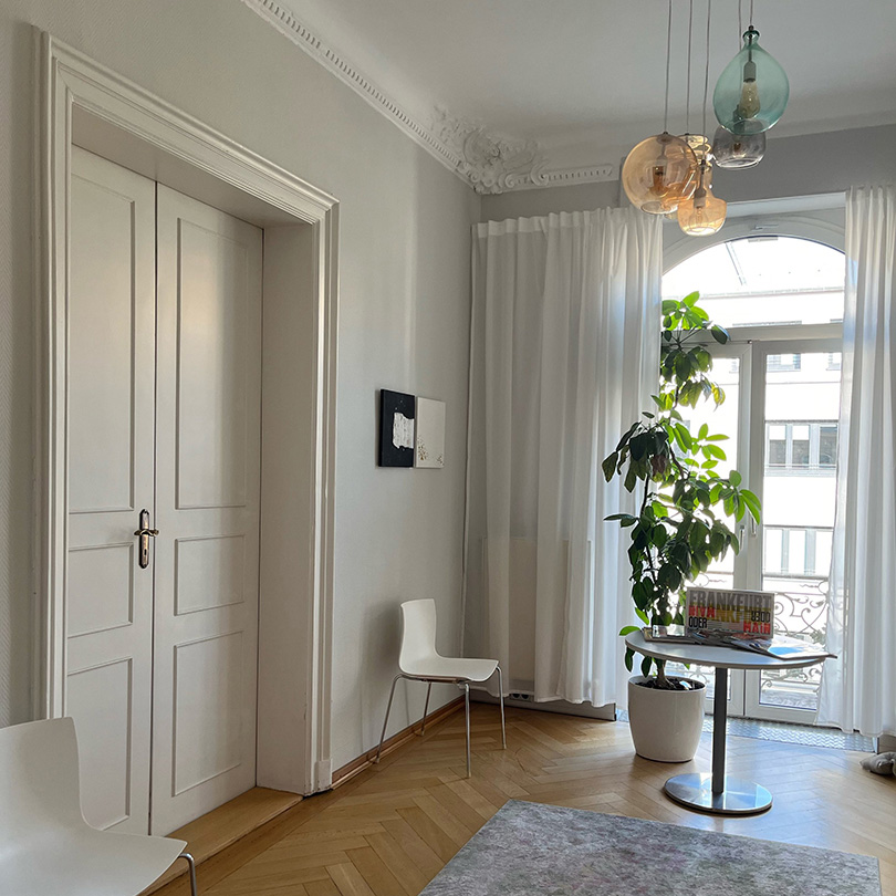 Wartebereich der Privatpraxis für Psychotherapie: ein heller Raum in einem stilvollen Altbau mit Stuckdecken, Holzparkett und weißen, modernen Möbeln.