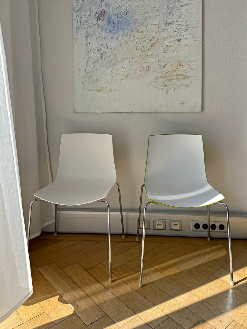 Wartebereich der Privatpraxis für Psychotherapie: ein Raum mit zwei Stühlen, Parkettboden und Kunst an der Wand. Durch einen leichten Vorhang fällt Sonnenlicht in das Zimmer.